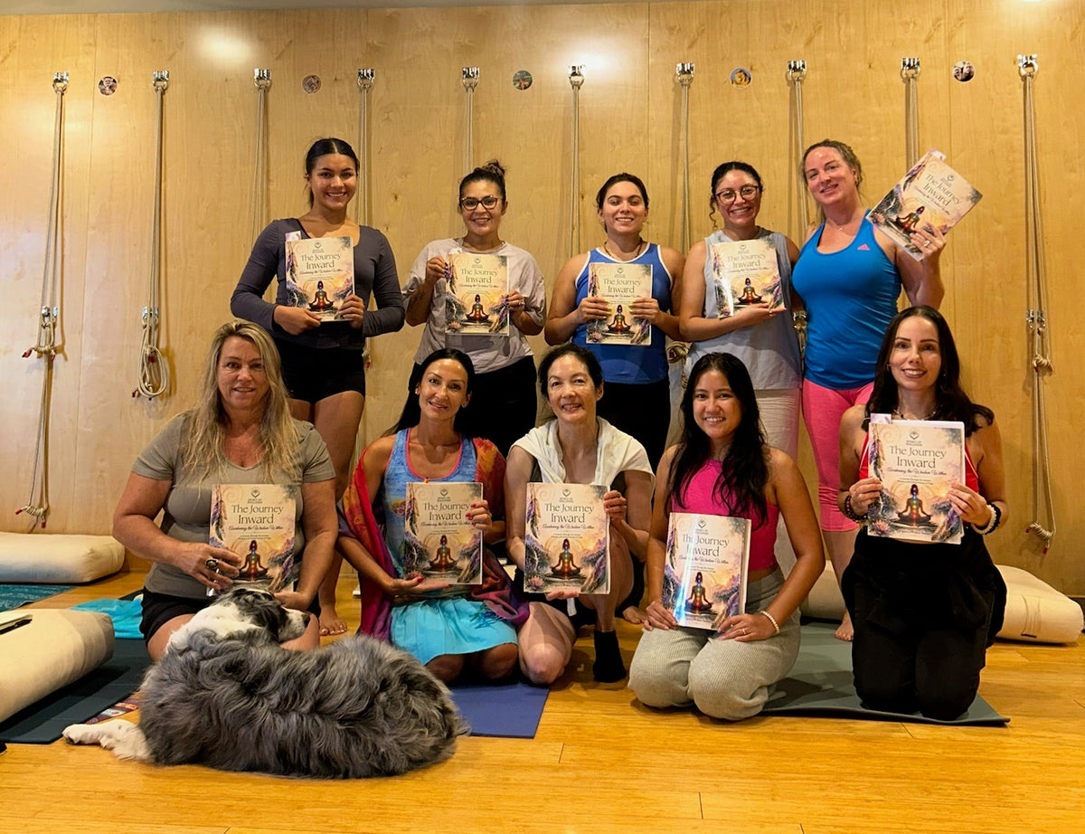 A group of nine women posing together in a yoga studio, each holding a copy of the book 'The Journey Inward'. A dog is lying on a yoga mat on the floor in the foreground.